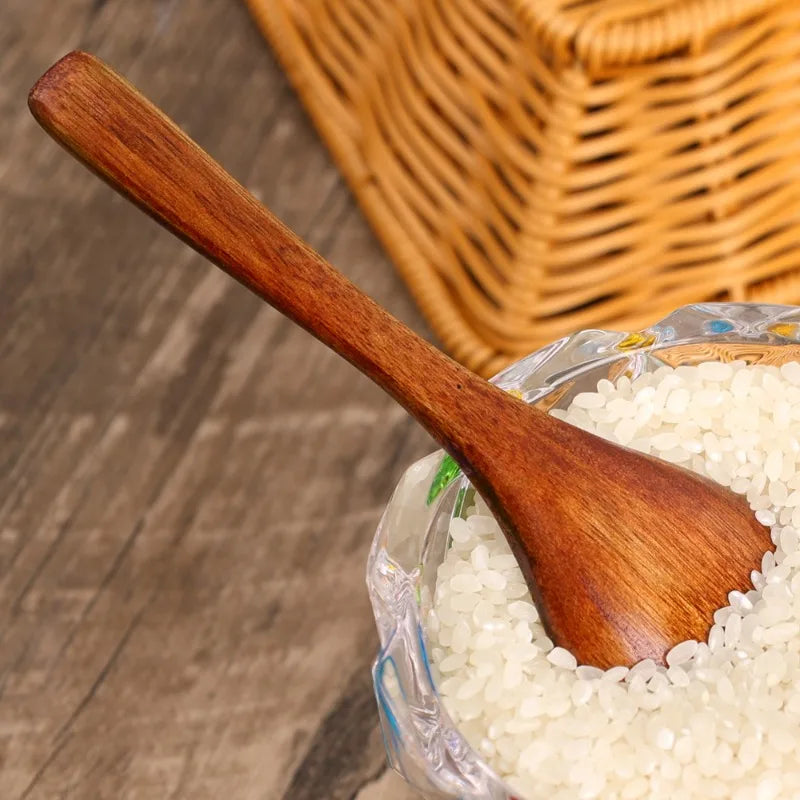 A wooden serving spoon is scooping white rice from a glass bowl, set against a blurred background of a woven basket.