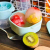 A bowl of colorful fruit spheres, including pink and yellow, next to a kiwi slice on a wooden table.