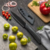 A set of three stainless steel utility knives resting on a dark surface with fresh produce and a wooden cutting board.