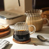 A handcrafted glass milk pot sits on a coaster next to a small spoon and bread on a white surface.