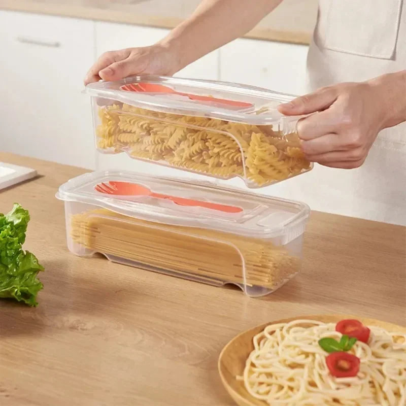 A hand is holding a clear plastic container filled with dry noodles, next to a larger container of similar noodles on a woode