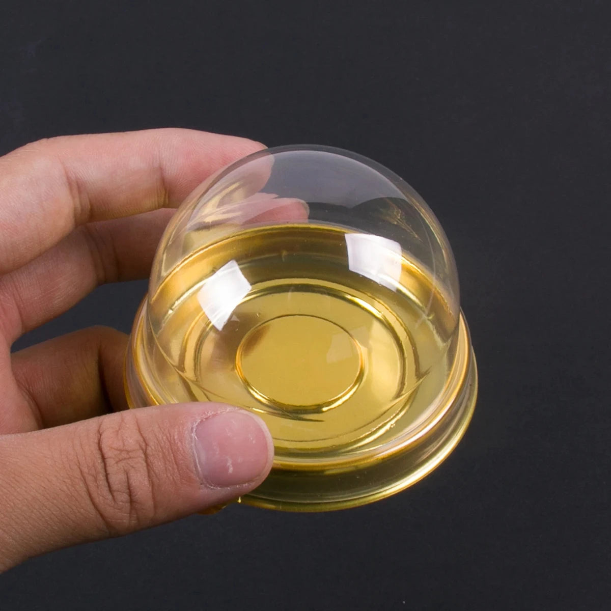 A hand holds a small, transparent glass cake box filled with golden-yellow liquid or oil.