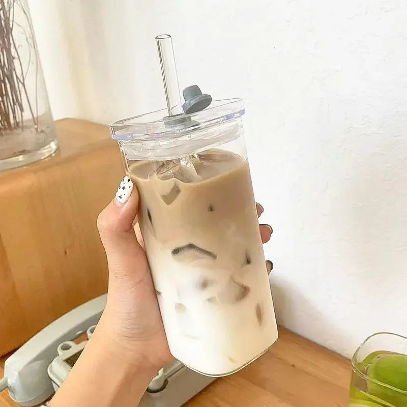 A hand holds a square heat resistant glass drink cup with lid and straw containing iced latte.