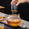 A hand pours amber liquid from a glass teapot into a cup, showcasing the hand-hammered glass design.