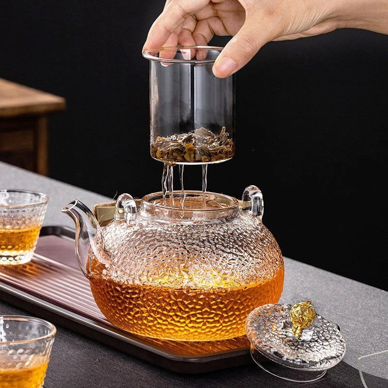 A hand pours amber liquid from a glass teapot into a cup, showcasing the hand-hammered glass design.