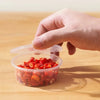 A hand is holding a clear plastic sauce cup filled with red chili flakes on a wooden surface.