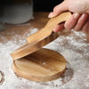 A hand is pressing a round wooden dumpling press tool onto a larger wooden base on a dusted surface.