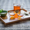 A wooden tray holds a stainless steel tea ball, a glass teapot, and tea leaves on a checkered cloth.