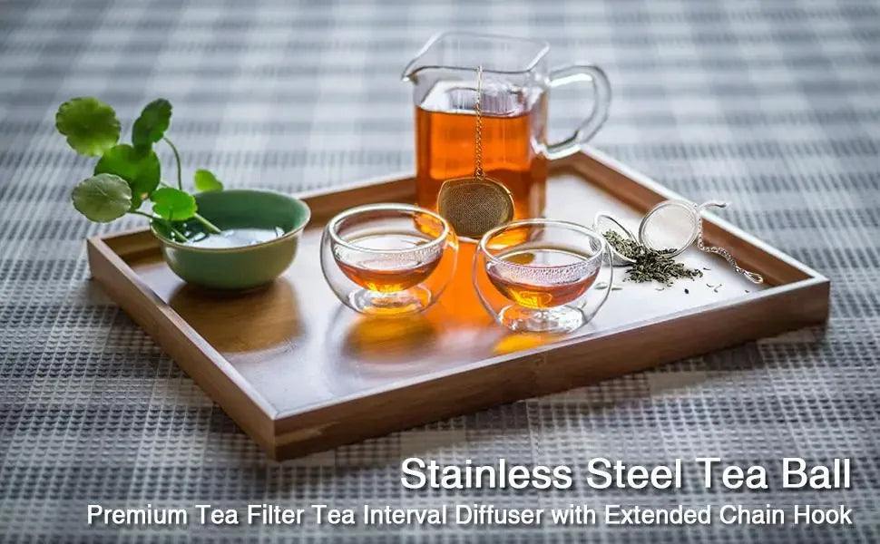 A wooden tray holds a stainless steel tea ball, a glass teapot, and tea leaves on a checkered cloth.