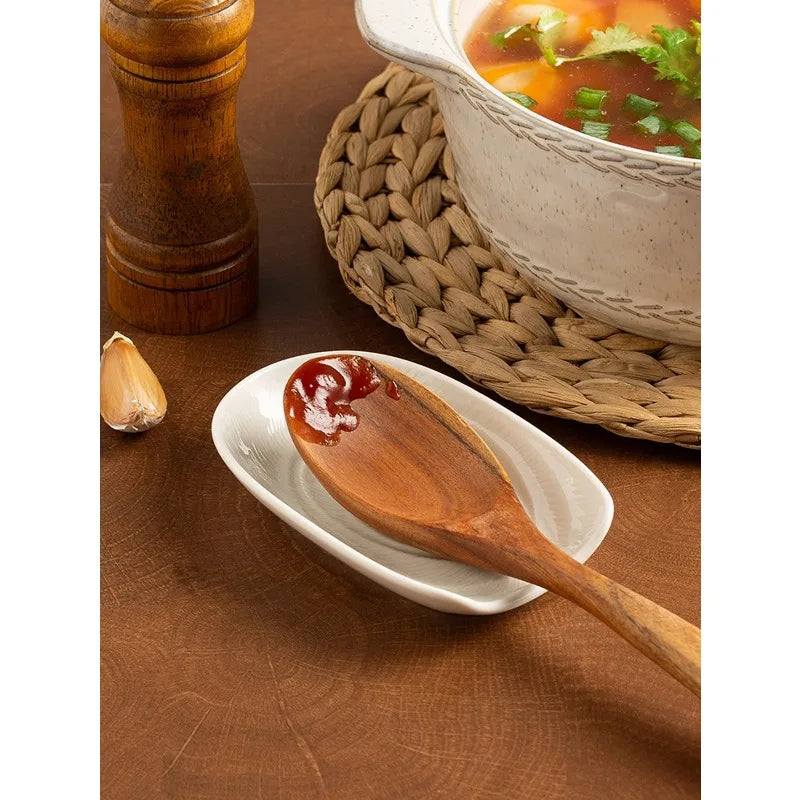 A wooden spoon rests on a white plate next to a ceramic bowl filled with soup, set against a woven mat.