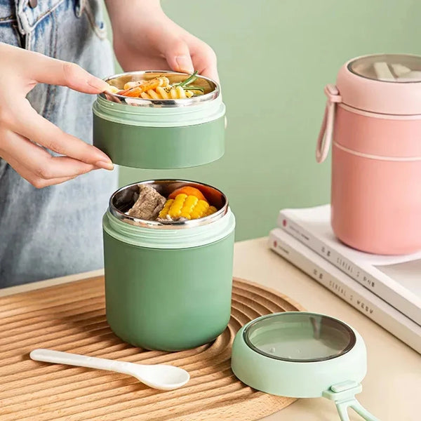 A hand is holding a green insulated stainless steel thermal food jar filled with soup, next to a wooden spoon and another jar