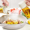 A hand is dispensing olive oil from a white spray dispenser into a pan containing meat and corn.