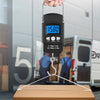 A hand holds a black portable digital hanging scale displaying 15.80, positioned above a cardboard box.