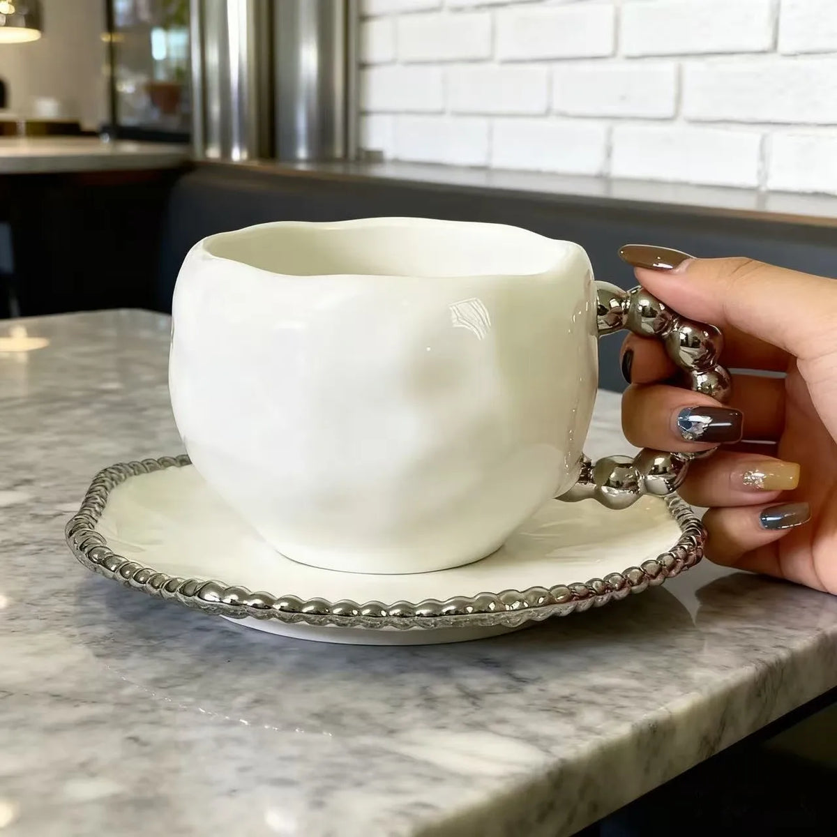 Une main tient un service de thé en céramique blanche avec des accents argentés sur une table en marbre.