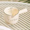 A white ceramic coffee cup with volume markings is shown on a striped surface.