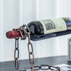 A dark glass bottle of wine is suspended by a metal chain, displayed on a white surface.