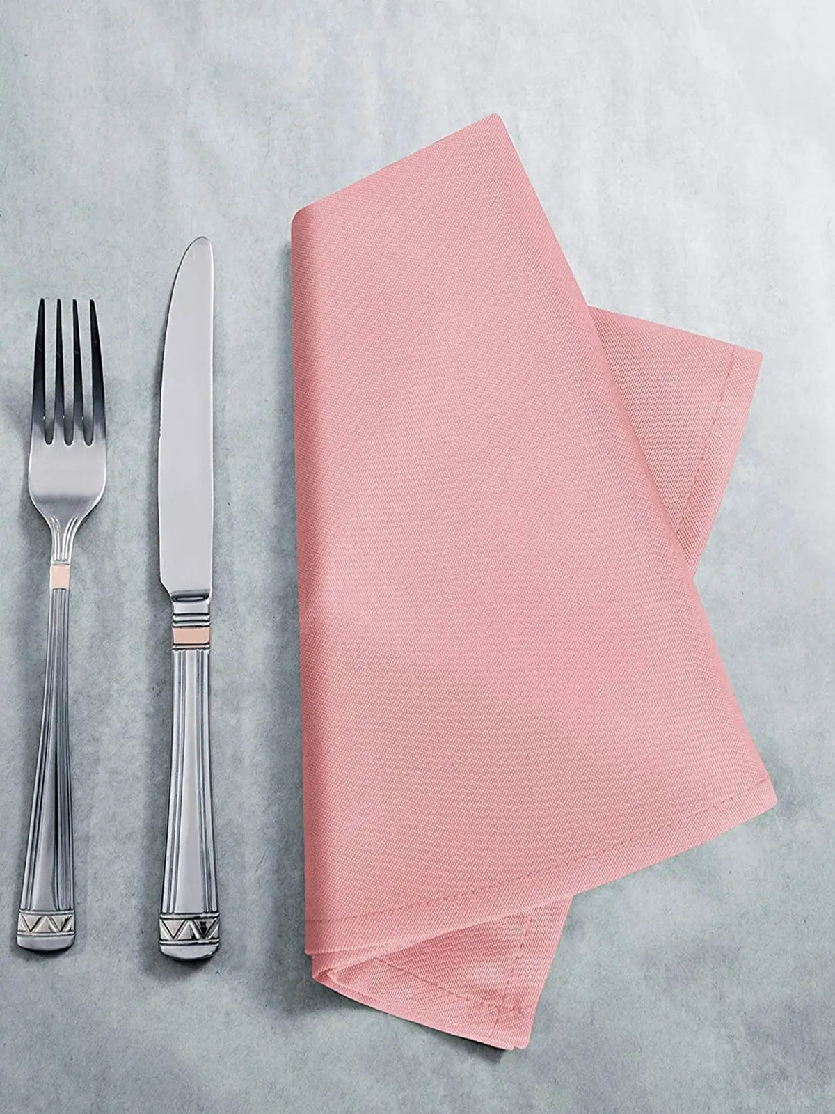 A stack of pink reusable weave tablecloth napkins next to a silver fork and knife on a light gray surface.