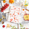 A spiral-bound recipe book with handwritten recipes and illustrations, surrounded by fresh ingredients like tomatoes and appl