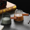 Una taza medidora de espresso de vidrio preciso con mango de madera sobre una superficie oscura junto a un libro.