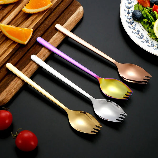 A set of two stainless steel forks and a spork, featuring gold and purple finishes, placed on a wooden board with fruit.