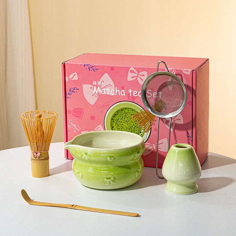 A pink box containing a Japanese matcha tool set with a ceramic bowl, bamboo whisk, and strainer on a white surface.