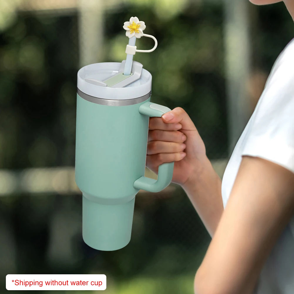 A hand holds a light green tumbler with a flower straw cap, set against a blurred outdoor background.