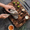 Hands slice a loaf of bread on a wooden cutting board with fresh limes and herbs.