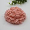 A pink silicone flower candle mold with layered petals resting on a white surface, with green foliage in the blurred backgrou