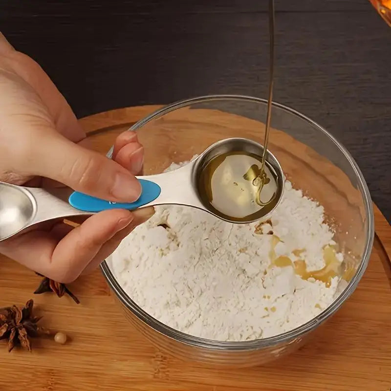 A hand pours oil from a stainless steel measuring spoon into a glass bowl containing white powder.