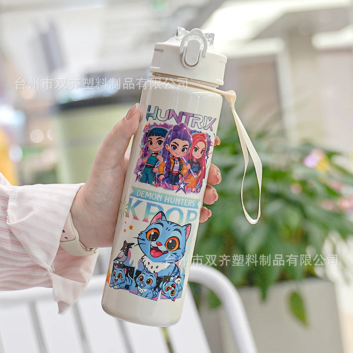 A hand holds a white insulated water cup featuring a cartoon print of demon hunters.