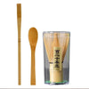 A glass container holding a set of bamboo and ceramic matcha tea tools, including a bamboo whisk and a bamboo spoon.