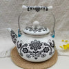 A white enamel teapot decorated with black floral and animal patterns sits on a woven coaster.
