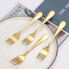 A set of six gold-toned stainless steel dessert forks arranged on a white plate.