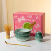 A pink box containing a Japanese matcha tea set with a ceramic bowl and whisk on a white surface.