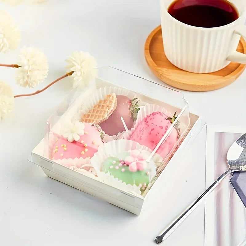 A clear square box filled with pink and green macarons and strawberries, set on a white surface.