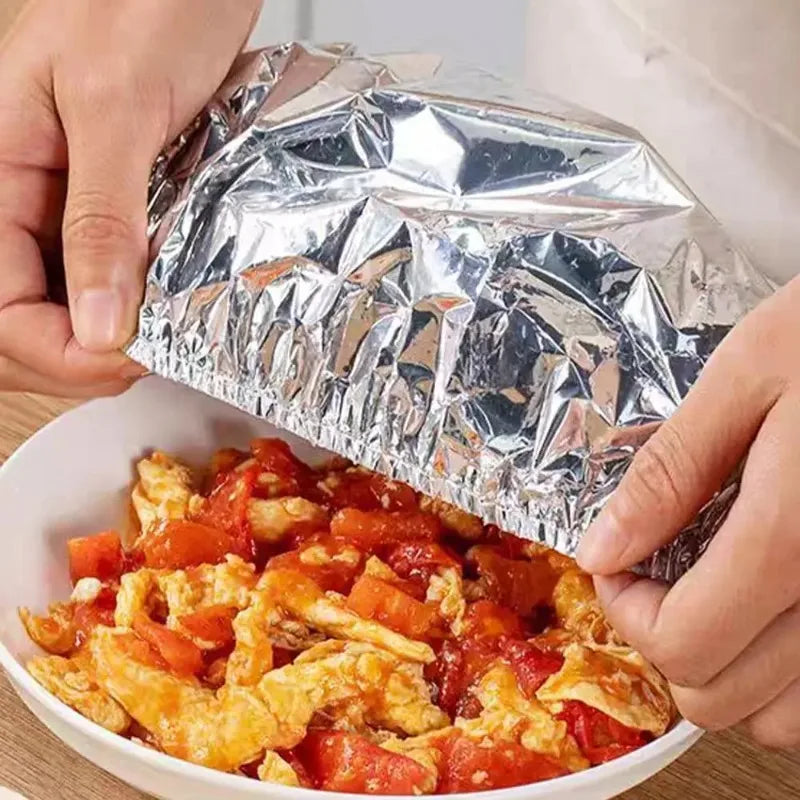 Hands are opening a package of aluminum foil food covers to reveal scrambled eggs in a white bowl.