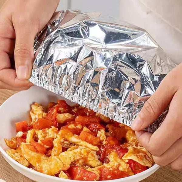 Hands are opening a package of aluminum foil food covers to reveal scrambled eggs in a white bowl.