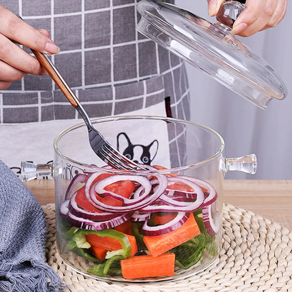Una mano usa un tenedor para remover verduras frescas como tomates y pepinos en una olla de vidrio transparente.