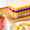 A stack of yellow and purple silicone ice cube trays on a wooden board next to a glass of pink fruit puree.