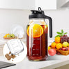 Botella de vidrio con asa negra llena de jugo rojo y rodajas de naranja sobre una encimera blanca.