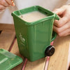A hand is pouring a light brown liquid into a green novelty trash can designed like a coffee mug.