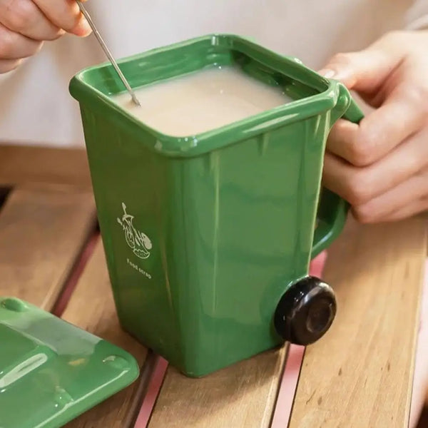A hand is pouring a light brown liquid into a green novelty trash can designed like a coffee mug.
