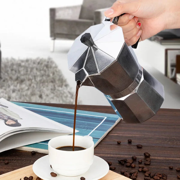 Eine Hand gießt dunken Kaffee aus einer italienischen Aluminium-Moka-Kanne in eine weiße Tasse auf einem Holztablett.