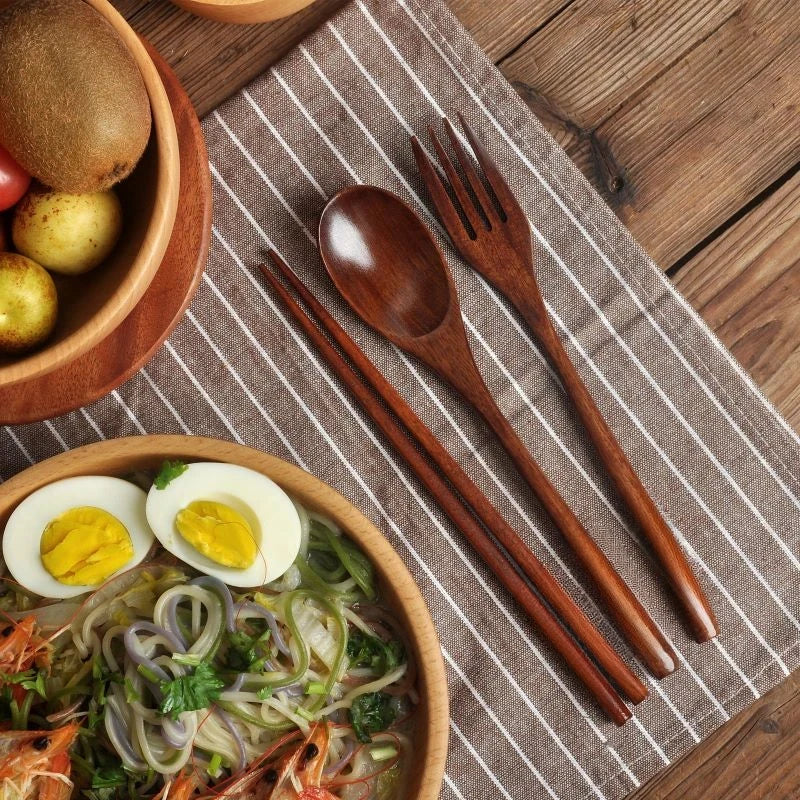 A natural wooden cutlery set including a fork, spoon, and chopsticks is displayed on a striped napkin with food.