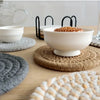 A set of four woven absorbent coasters displayed on a wooden table with white bowls and natural fiber mats.