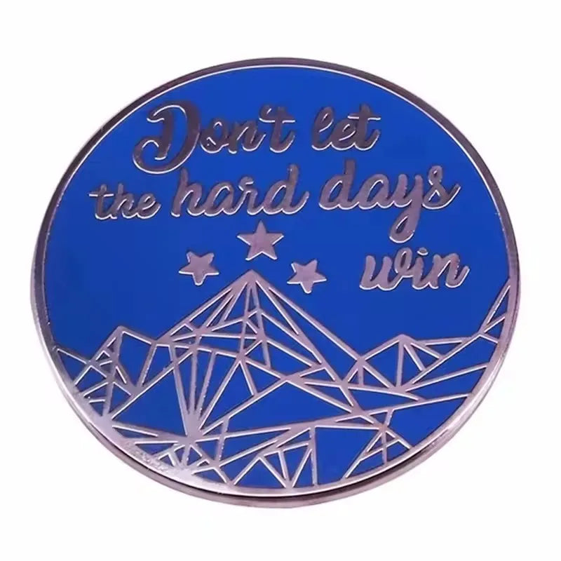 A blue enamel pin featuring the text Don't let the hard days win and a geometric mountain design.