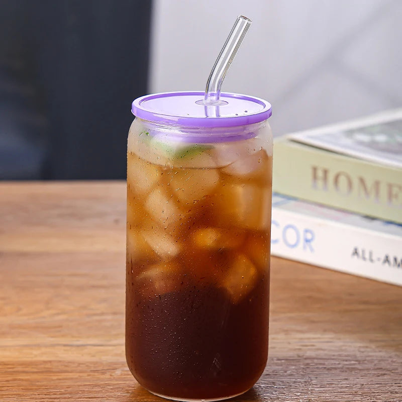 A glass cup filled with iced tea or a dark beverage, topped with a purple bamboo lid and straw, resting on a wooden surface.