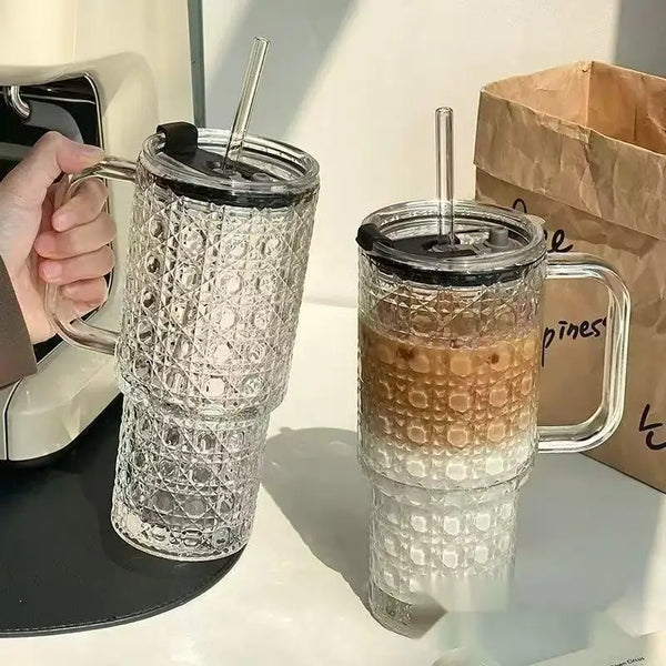 Klare, strukturierte Glas-Tasse mit Henkel, Deckel und Strohhalm neben einem braunen Papiertüte.