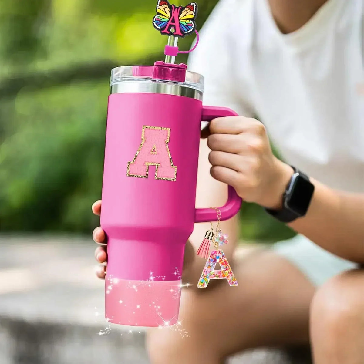 A pink tumbler with a glitter gold letter A and an attached butterfly straw cover is held by a person outdoors.