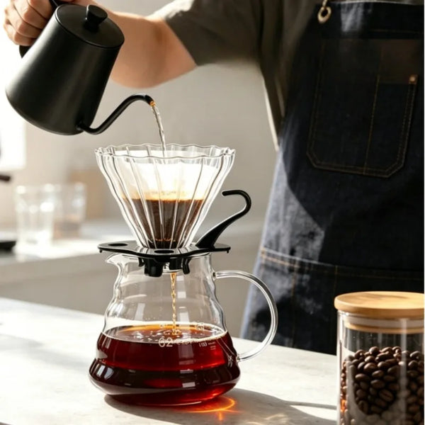 A hand pours brewed coffee from a black manual pour over dripper into a glass carafe.